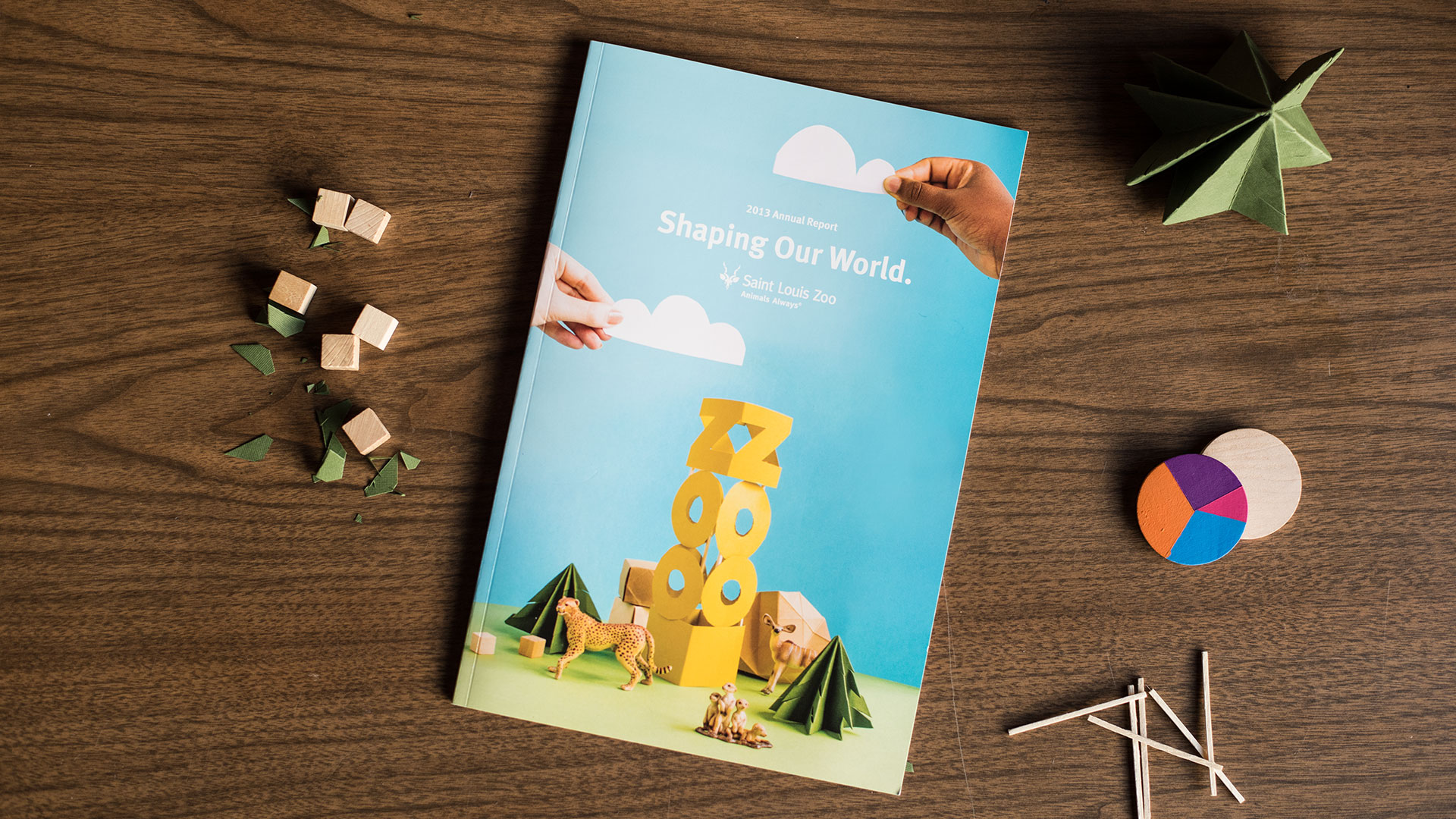 Saint Louis Zoo 2013 Annual Report cover "Shaping Our World" on a wood desk with paper-craft props