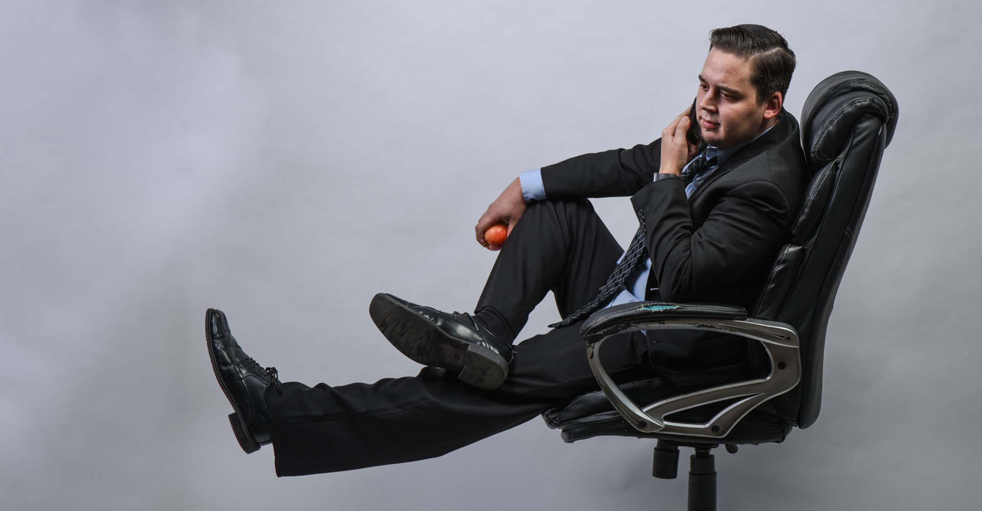 Drew Douglass wearing a suit, legs crossed in a computer chair purchased at a club store, holding a phone up to his ear in one hand, and an apple in the other hand. Also - his legs are resting on an invisible box.