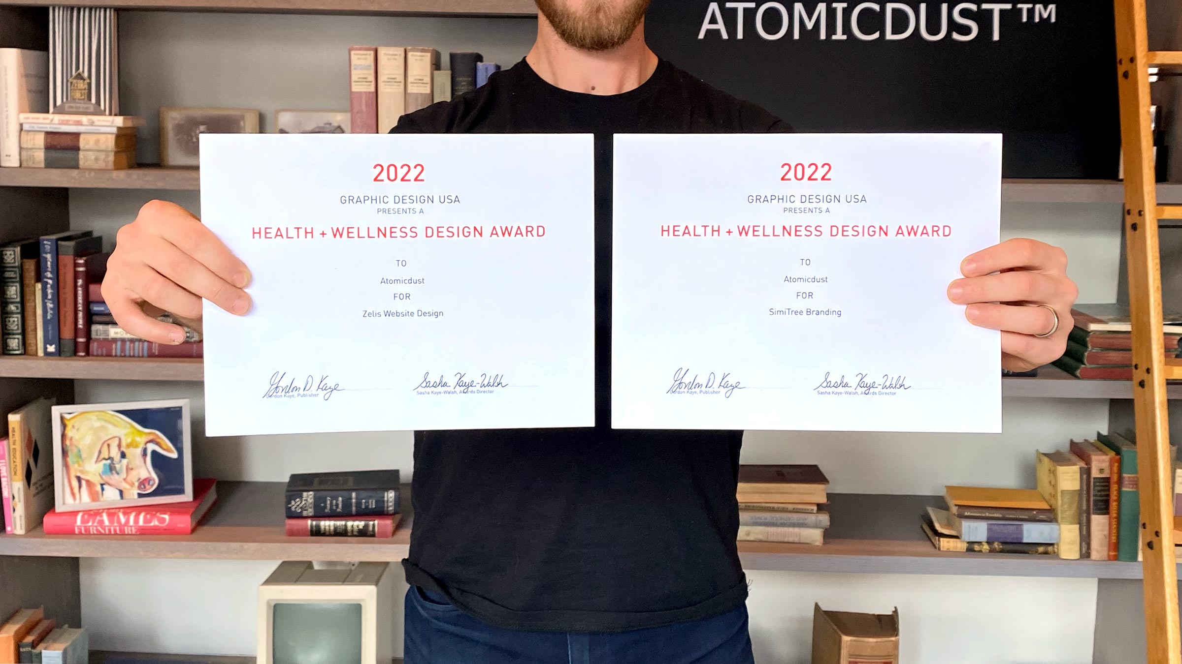 An Atomicdust team member holds two Graphic Design USA Health and Wellness 2022 winners certificates
