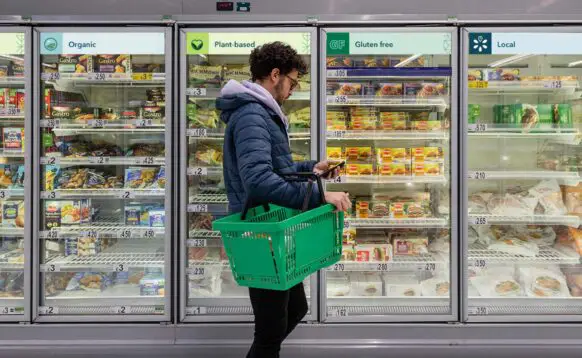 A person walks past the freezer aisle of the grocery store with Eatwell branding on signage