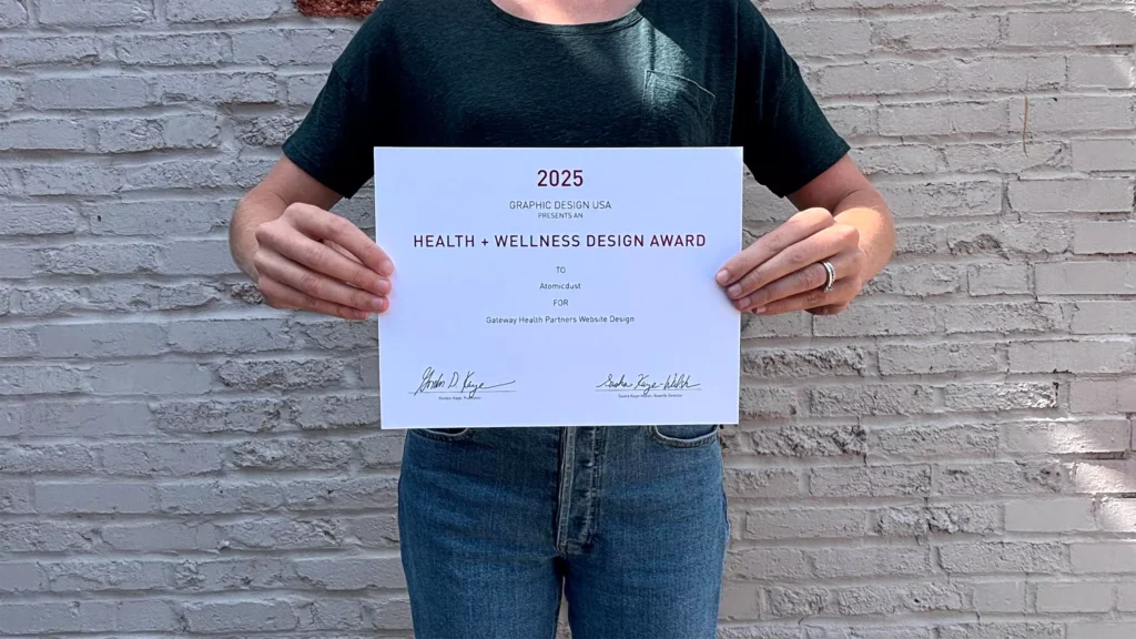 An Atomicdust team member holds a 2025 Graphic Design USA Health + Wellness Awards certificate