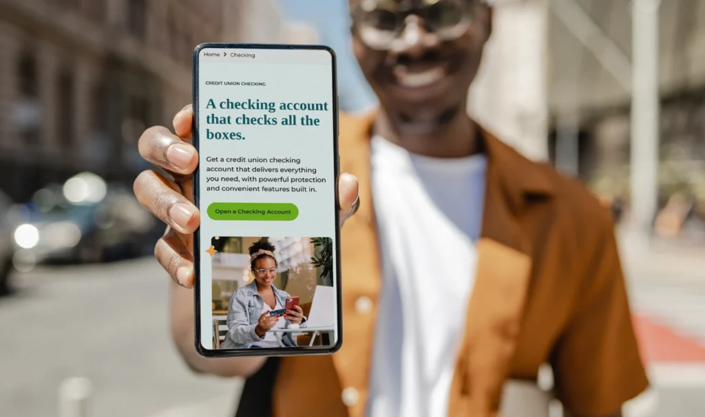 A smiling man holding a smartphone displaying the Vantage Credit Union mobile checking account page on a city street