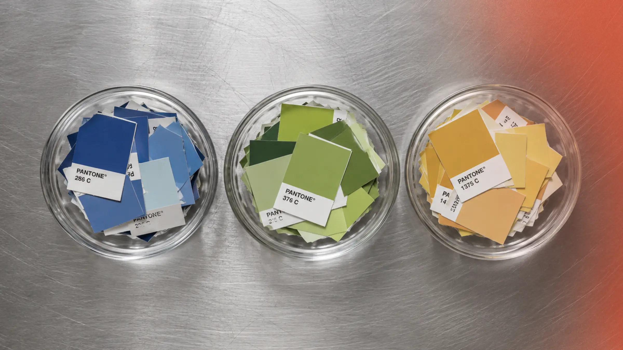 Three glass prep bowls on a stainless steel counter, viewed from above, each filled with Pantone color chips in blues, greens and golden yellows, arranged like ingredients ready for mise en place.